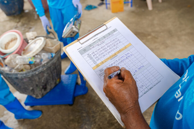 A worker writes in a data sheet as two other workers weigh a basket of trash