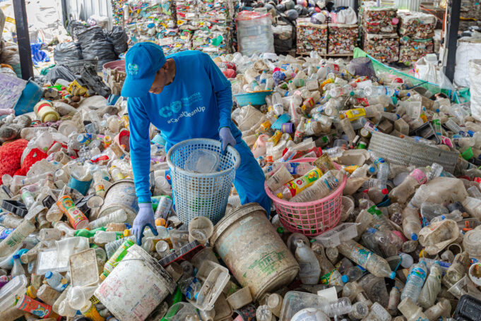 A worker sorts plastics by hand, surrounded by hundreds of plastic bottles