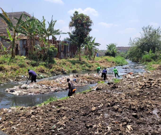 In the foreground, a muddy riverbank. 5 volunteers wearing green are in the river removing trash. The river flows through the photo from left to right. An island of garbage is formed in the middle of the river. Across the river, the river bank is sloped and covered in grass, with tropical trees growing around a small wooden house.