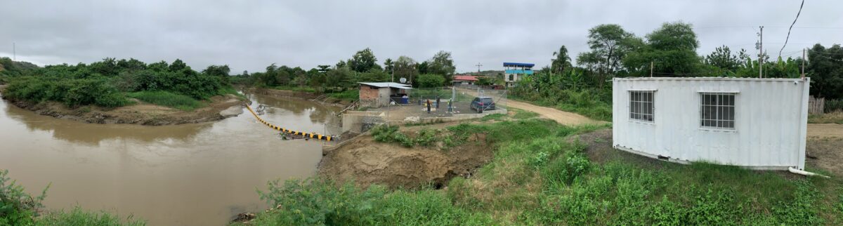Panoramic view of a brown, muddy river and the river bank. The river is flowing from the upper left of the image, to the middle, and turning back out of view on the lower left. A yellow and black floating boom crosses the river at the bend, and on the right side of the river, the boom leads to a concrete structure with a flat area at which trash is collected from the river. In the back, there are three structures in the distance. In the front right, a white shed with two windows.