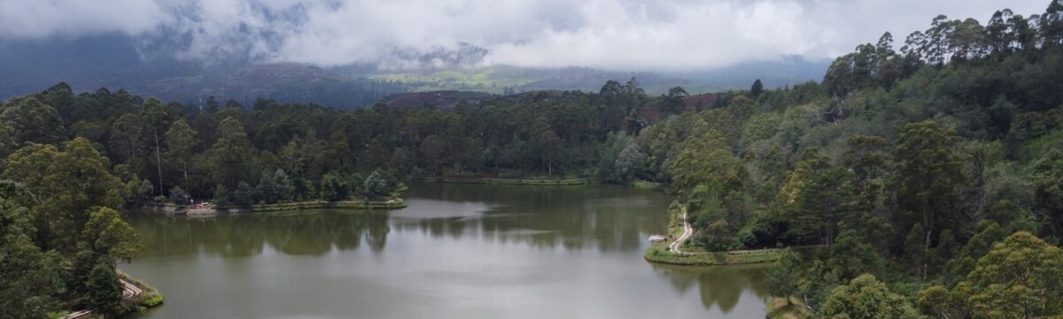 A wide, peaceful river meanders through the middle of the frame. Dense green vegetation covers the riverbanks up to the waterline. In the distance, low clouds and mist hang over hills.