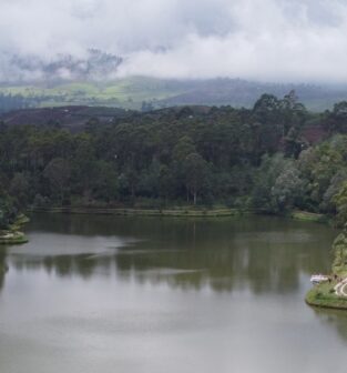 A wide, peaceful river meanders through the middle of the frame. Dense green vegetation covers the riverbanks up to the waterline. In the distance, low clouds and mist hang over hills.