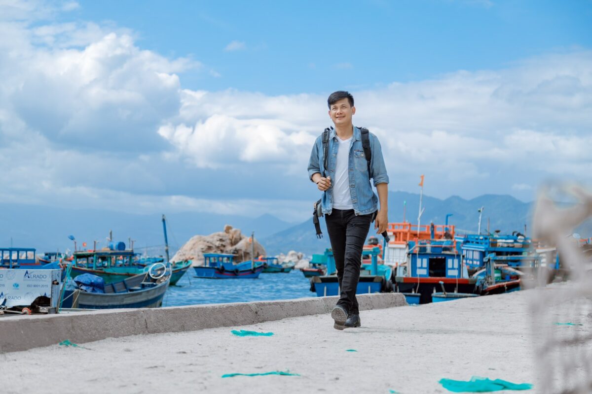 A man with short black hair in black jeans, a white t-shirt, and a blue denim jacket wears a backpack an walks towards the camera on a cement pier. Behind him, small fishing boats loaded with nets and traps float on aqua blue water with big white clouds dotting a blue sky.