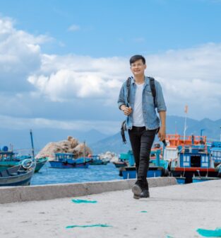 A man with short black hair in black jeans, a white t-shirt, and a blue denim jacket wears a backpack an walks towards the camera on a cement pier. Behind him, small fishing boats loaded with nets and traps float on aqua blue water with big white clouds dotting a blue sky.