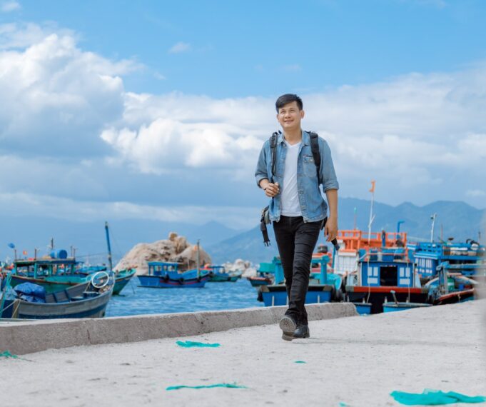 A man with short black hair in black jeans, a white t-shirt, and a blue denim jacket wears a backpack an walks towards the camera on a cement pier. Behind him, small fishing boats loaded with nets and traps float on aqua blue water with big white clouds dotting a blue sky.