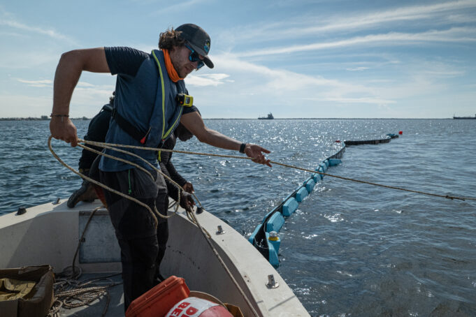 A man stands on a boat in the ocean, in action pulling a rope towards him. He stands on the bow, the photo is taken from the boat. Extending from the boat in the background is a floating blue chain link barrier, which is being installed by the person in the photo to anchor points.