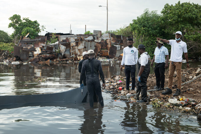 Six people gather at the water's edge within a gully. Two are in the the water, the other four stand on land. The water is dark and polluted, the people in the water wear full wetsuits. On shore, there is trash all around. Across the water in the background, pieces of scrap metal are pieced together to create a fence.
