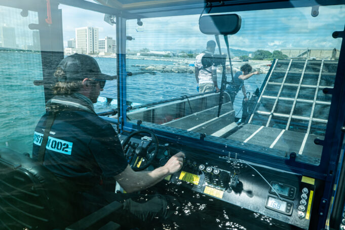 A person, photo taken from behind, sits at the helm of a boat. The boat is enclosed by a clear window, and through the window the front of the boat can be seen. The front of the boat is a mechanical conveyor belt, currently tilted upward on hinges. Two people outside on the front of the boat work on the conveyor. In the distance, the harbor and buildings.