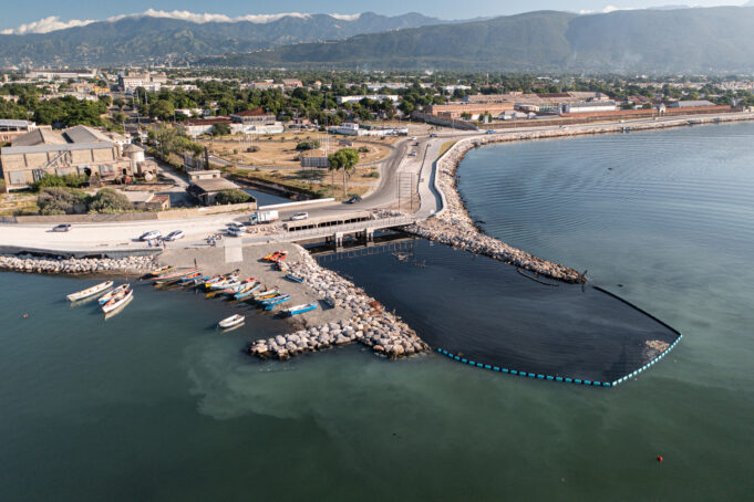 An aerial image of the coastline of Kingston Harbour. A drainage gully empties through coastal infrastructure, and the floating blue barrier extends across the opening of where the gully meets the ocean. In the distance, villages and mountains.