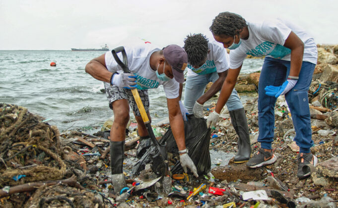 Three people work together to put waste into a garbage bag. They kneel down with gloves and shovels. They are surrounded by piles of waste, and stand at the edge of the water in the harbor with a ship and buoy in the distance.