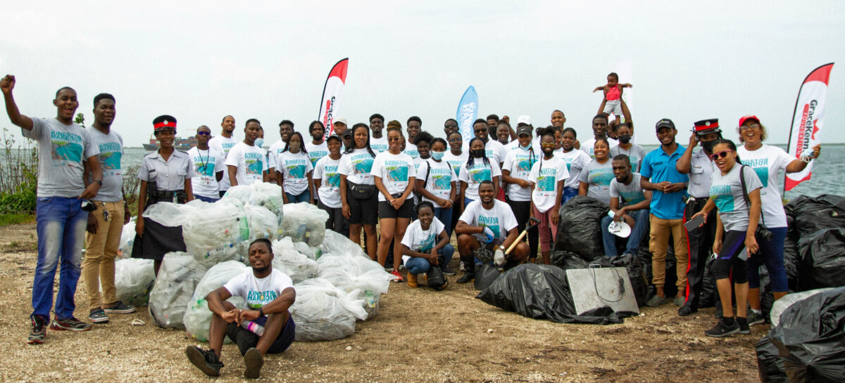 A large group of volunteers, roughly 50 people, gather in two lines on the beach for a photo. They have just completed a beach clean up, and are standing around the many large garbage bags filled with the waste they cleaned up.