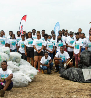 A large group of volunteers, roughly 50 people, gather in two lines on the beach for a photo. They have just completed a beach clean up, and are standing around the many large garbage bags filled with the waste they cleaned up.