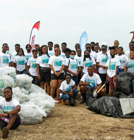 A large group of volunteers, roughly 50 people, gather in two lines on the beach for a photo. They have just completed a beach clean up, and are standing around the many large garbage bags filled with the waste they cleaned up.