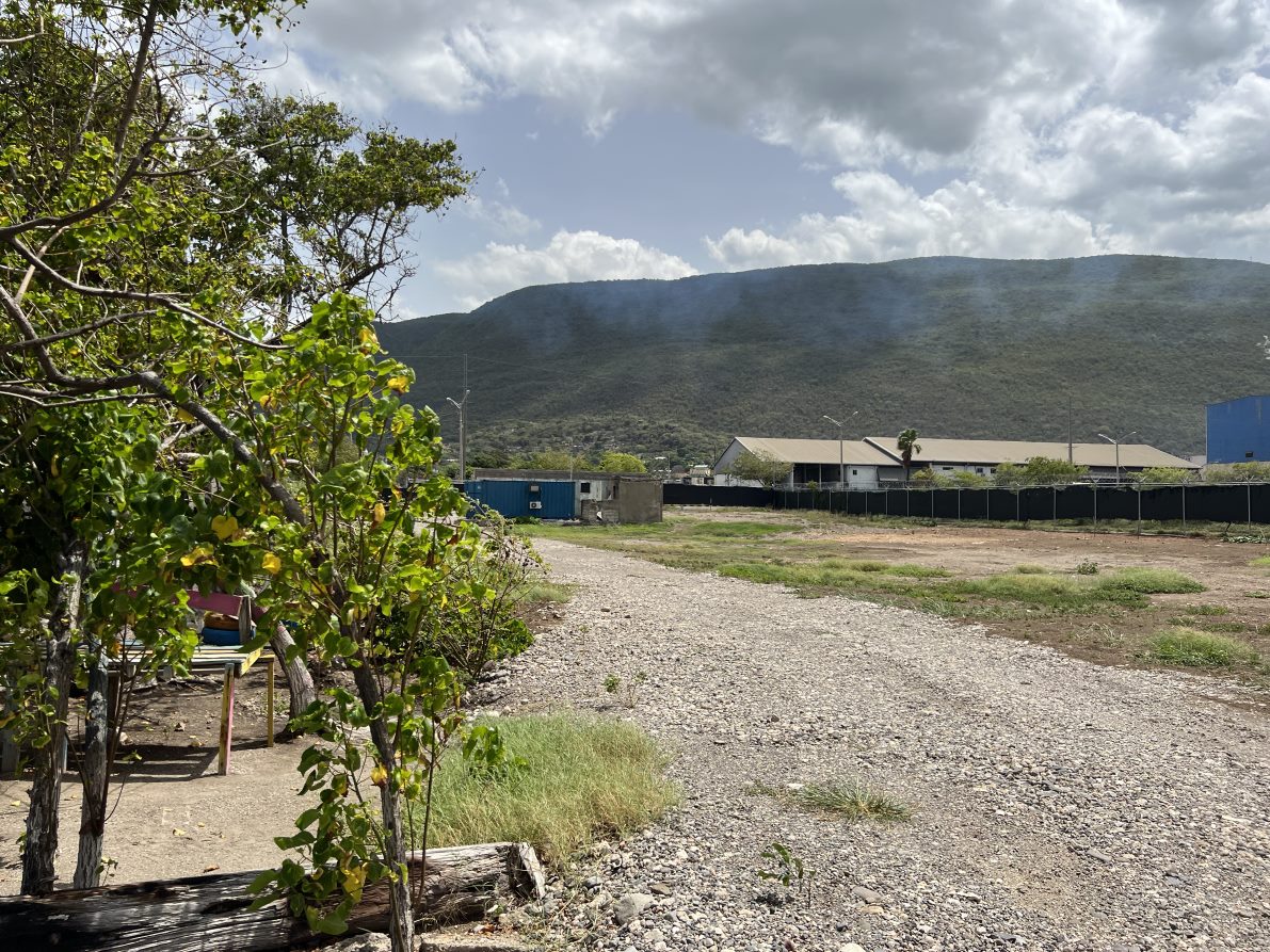 Small trees in the foreground on the left. In the center, a gravel driveway. To the right, open land is enclosed by a chain link fence with a large building behind and mountains in the distance.