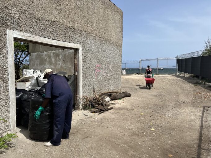 On the left, a concrete building with a wide open doorway. A person is holding a large trash bag in the doorway, and through the doorway many more trash bags are visible. To the right, a chain link fence surrounds the property, and in the distance is the ocean. In the center from a distance, a person approaches the doorway pushing a red wheelbarrow.