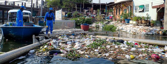 A mix of floating trash and water hyacinth plants accumulate between two extended metal boom arms and trap, floating in a narrow canal waterway. Behind the trap, tightly packed and colorful houses sit against the waters edge, buffered by a wall of sandbags. Next to the trap floats a small blue boat. A man in all blue and a face covering stands in the boat, looking at the trash. Another man, also in all blue, stands on the trap, looking down at the trash. Behind them, a concrete bridge spans the channel.