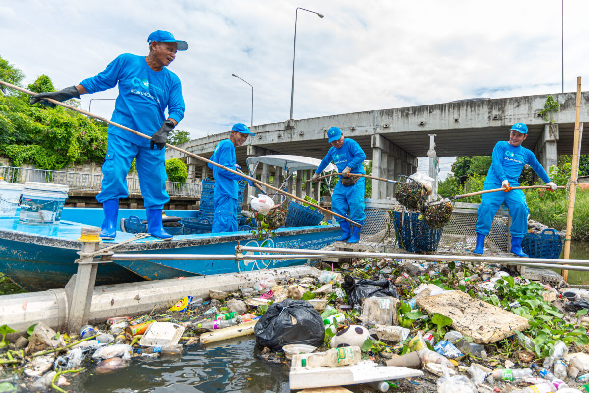 Four men in blue pants, long sleeve blue shirts, and blue hats weild bamboo poles with nets on the end. Three stand atop a small blue boat, another atop a metal trash trap floating next to the boat in a canal. The trap is full of floating garbage, and the men smile as they scoop the trash from the trap and put it in the boat. Behind them, a bridge spans the canal.