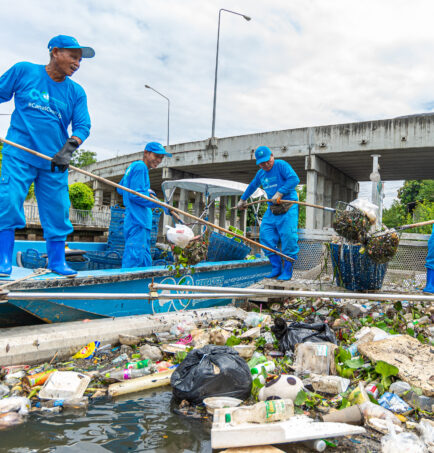 Four men in blue pants, long sleeve blue shirts, and blue hats weild bamboo poles with nets on the end. Three stand atop a small blue boat, another atop a metal trash trap floating next to the boat in a canal. The trap is full of floating garbage, and the men smile as they scoop the trash from the trap and put it in the boat. Behind them, a bridge spans the canal.