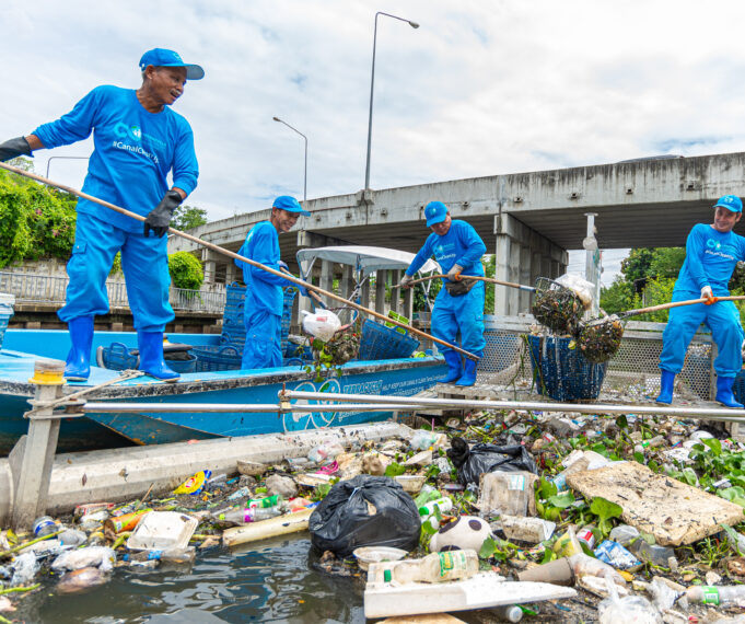 Four men in blue pants, long sleeve blue shirts, and blue hats weild bamboo poles with nets on the end. Three stand atop a small blue boat, another atop a metal trash trap floating next to the boat in a canal. The trap is full of floating garbage, and the men smile as they scoop the trash from the trap and put it in the boat. Behind them, a bridge spans the canal.